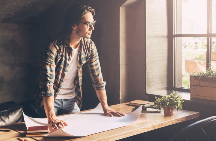 Man in plaid shirt leans over desk with large paper, looking out a sunlit window. Potted plants on desk, creating a contemplative mood.