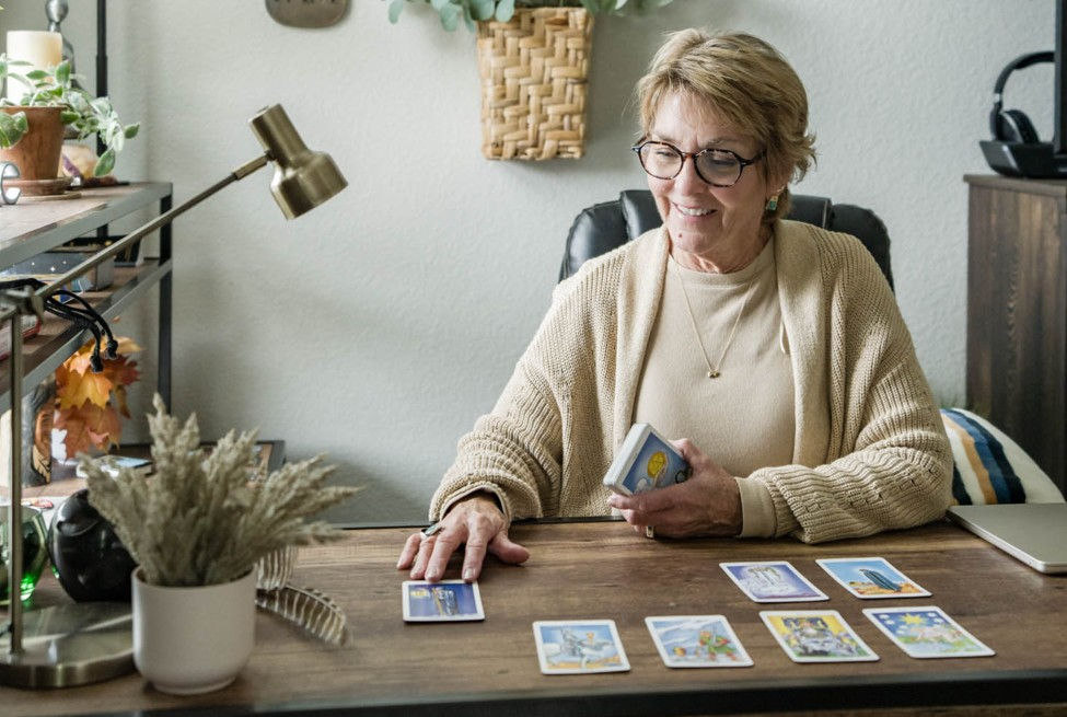 Smiling older woman reads tarot cards at a desk in a cozy home office, with several cards laid out before her.