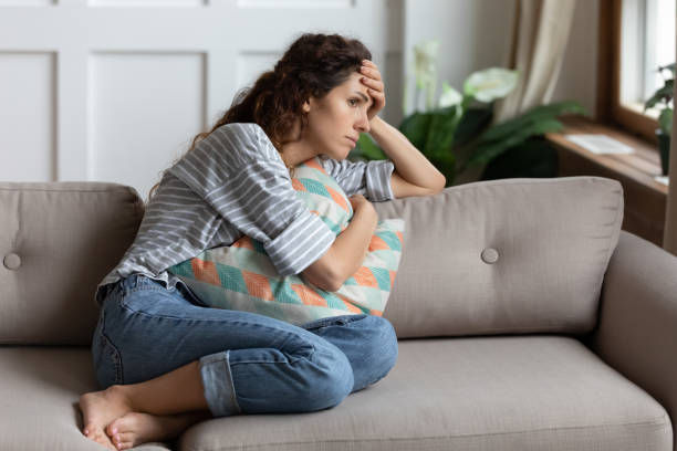 Frustrated young lady sitting on sofa, cuddling pillow, looking away at window.