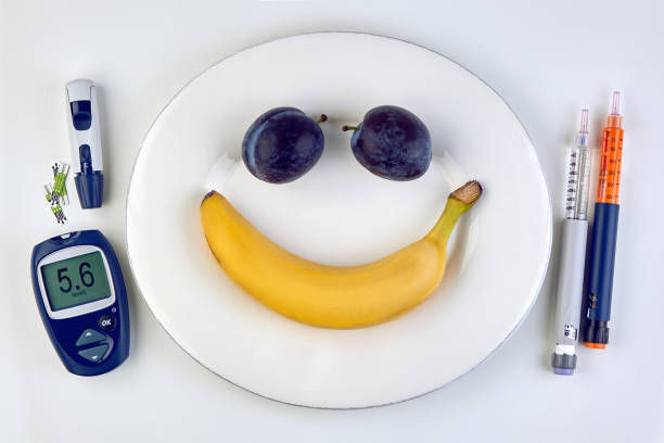 Banana and plums in the form of a smiling emoticon on a white plate, insulin syringe pens and a glucometer as cutlery on a white table.
