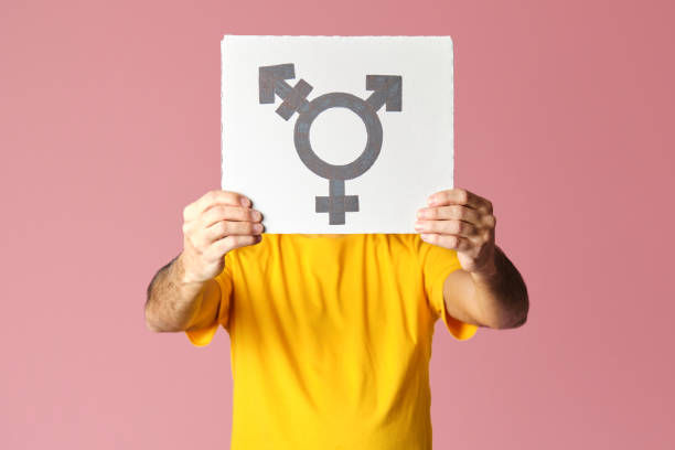 Man in yellow t-shirt holding a white sign covering his face with the symbol of the transgender in pink background.