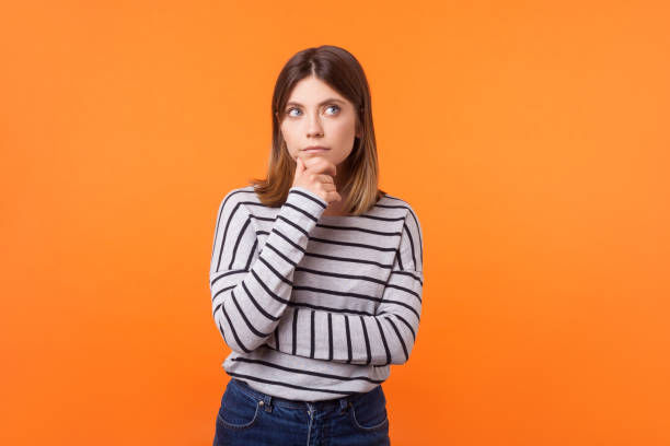 Portrait of pensive woman with brown hair in long sleeve striped shirt standing holding hand on chin and looking up